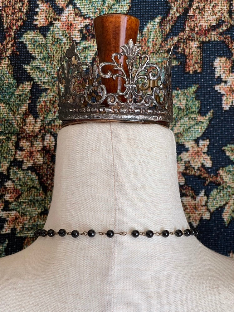 A historically inspired black beaded necklace with a bronze gothic medieval cross pendant is pictured on a mannequin in front of a floral tapestry.