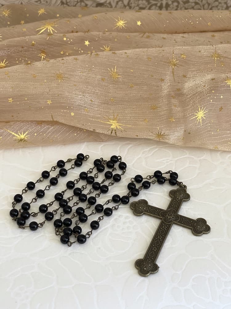 A historically inspired black beaded necklace with a bronze gothic medieval cross pendant is pictured on a floral puff with celestial fabric background..