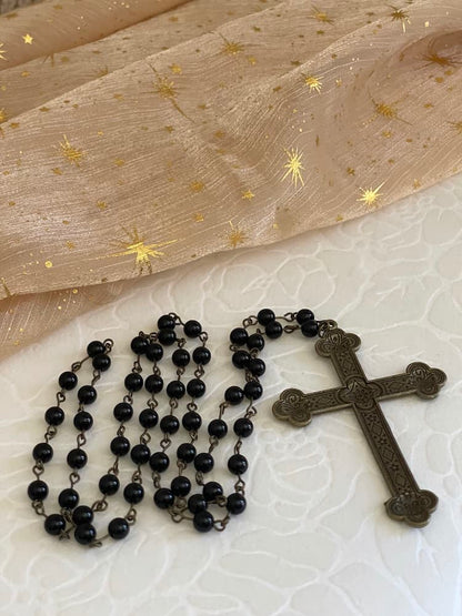 A historically inspired black beaded necklace with a bronze gothic medieval cross pendant is pictured on a floral puff with celestial fabric background..