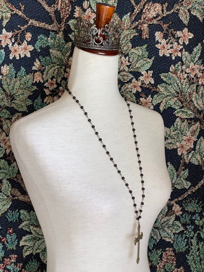 A historically inspired black beaded necklace with a bronze gothic medieval cross pendant is pictured on a mannequin in front of a floral tapestry.