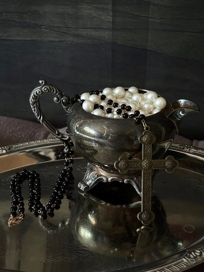 A historically inspired black beaded necklace with a bronze gothic medieval cross pendant is pictured on a mannequin in front of a cathedral backdrop.