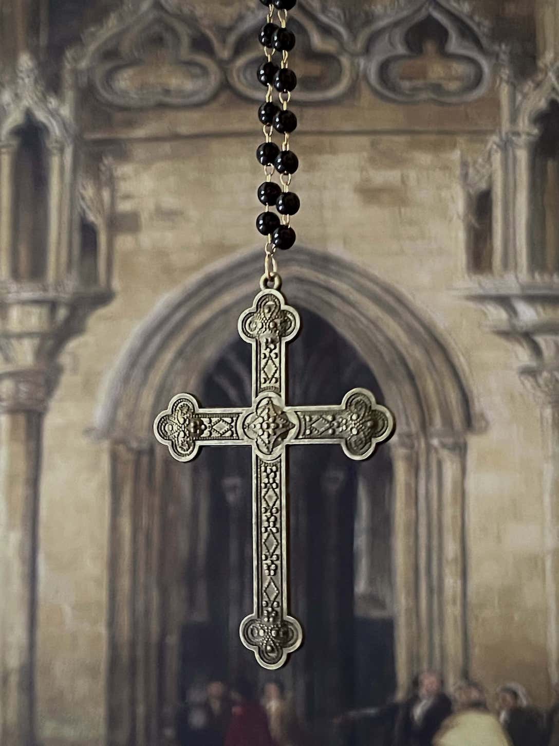 A historically inspired black beaded necklace with a bronze gothic medieval cross pendant is pictured on a mannequin in front of a cathedral backdrop.