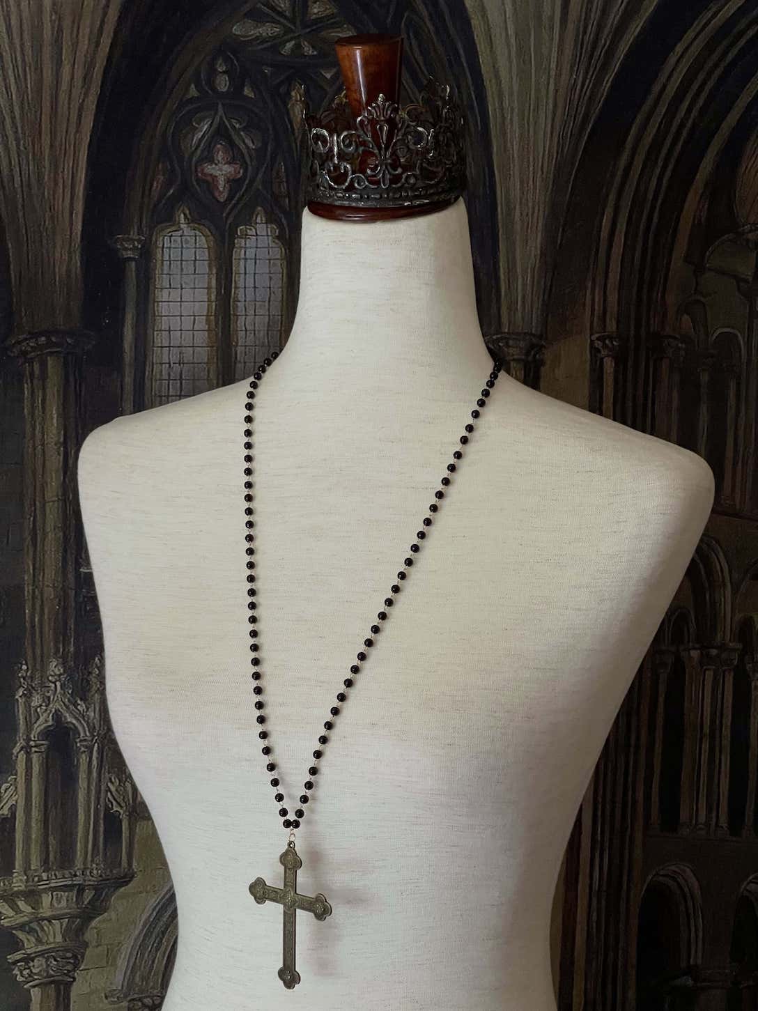 A historically inspired black beaded necklace with a bronze gothic medieval cross pendant is pictured on a mannequin in front of a cathedral backdrop.