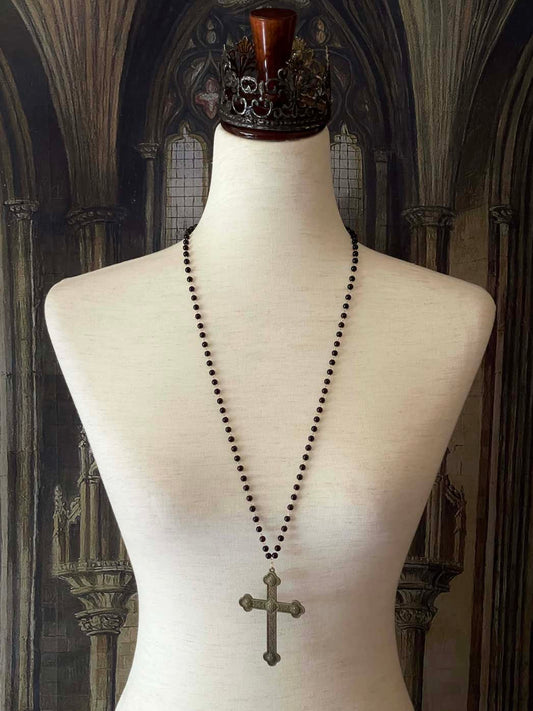 A historically inspired black beaded necklace with a bronze gothic medieval cross pendant is pictured on a mannequin in front of a cathedral backdrop.