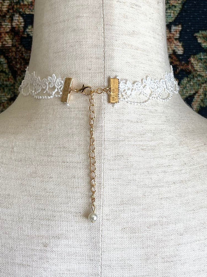 A Rococo or Regency era historically inspired pearl and floral lace choker necklace is pictured on a mannequin in front of a historic tapestry.