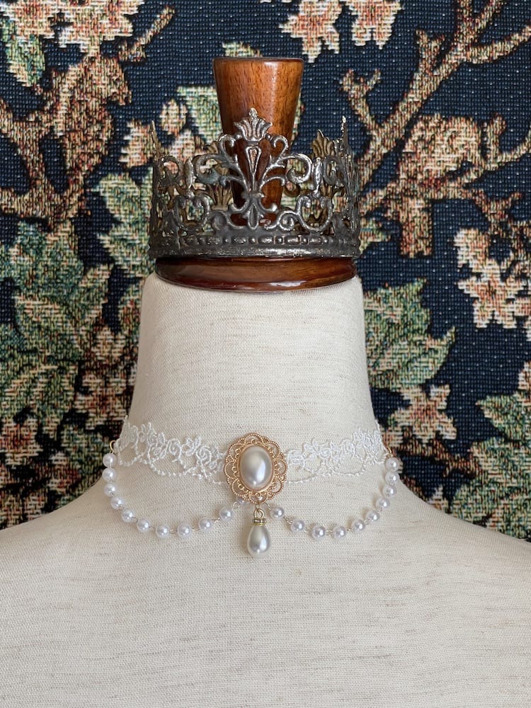 A Rococo or Regency era historically inspired pearl and floral lace choker necklace is pictured on a mannequin in front of a historic tapestry.