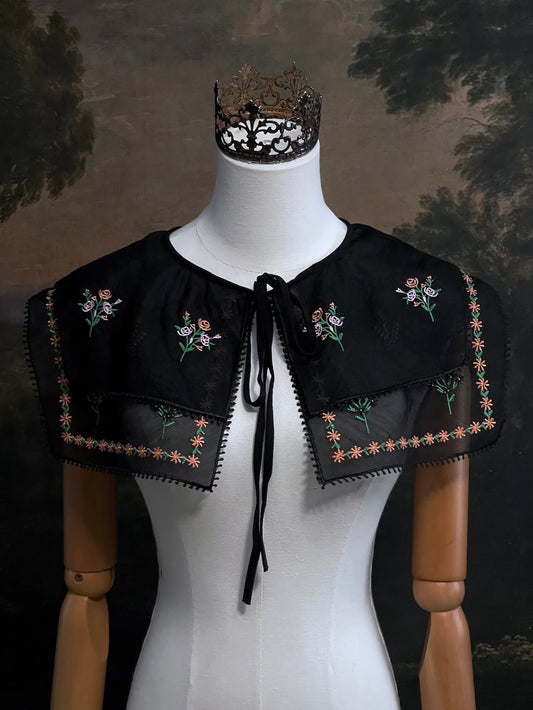 Mannequin wearing a black embroidered cape collar with floral designs against a dark art history painting background