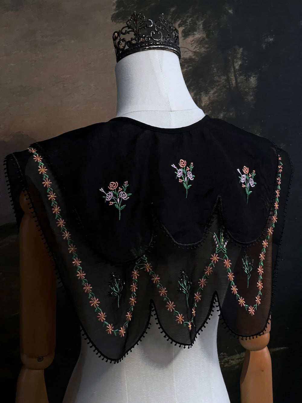Mannequin wearing a black embroidered cape collar with floral designs against a dark art history painting background