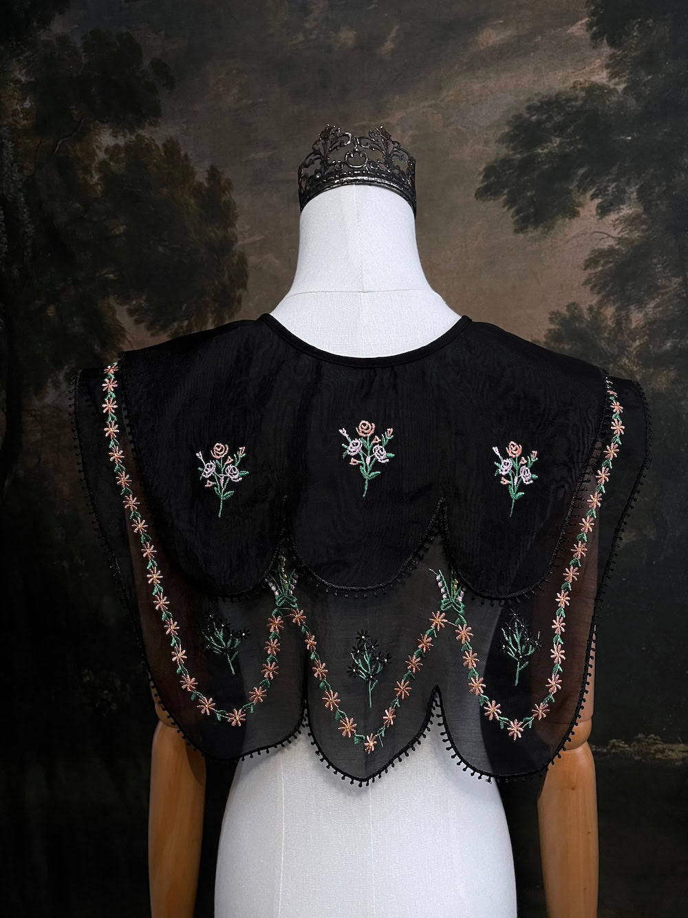 Mannequin wearing a black embroidered cape collar with floral designs against a dark art history painting background