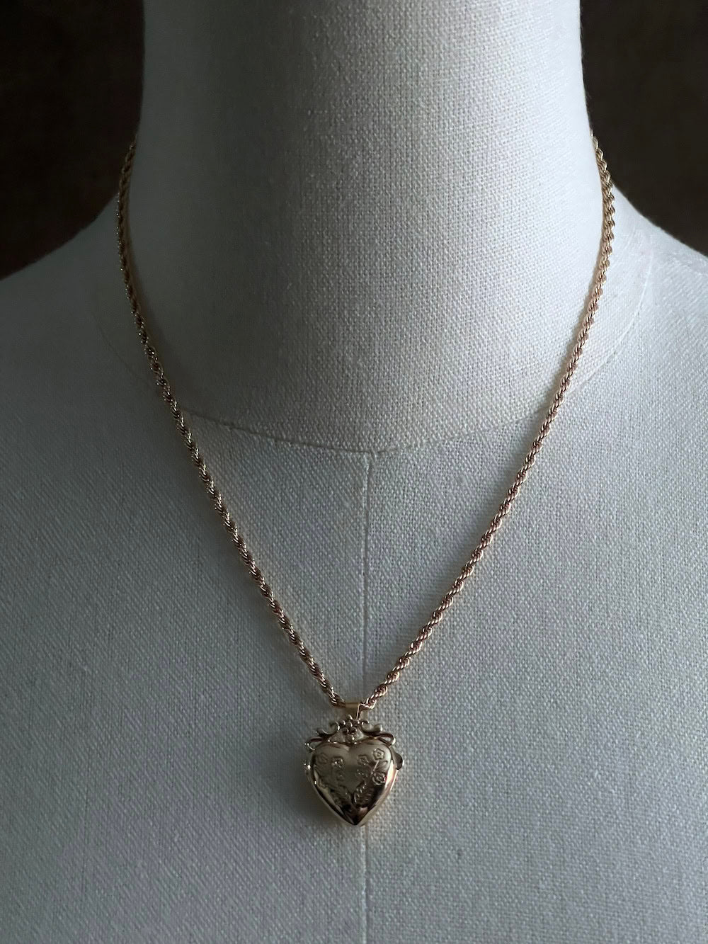 Mannequin wearing a gold twisted chain with a heart shaped locket pendant carved with historically inspired floral filigree