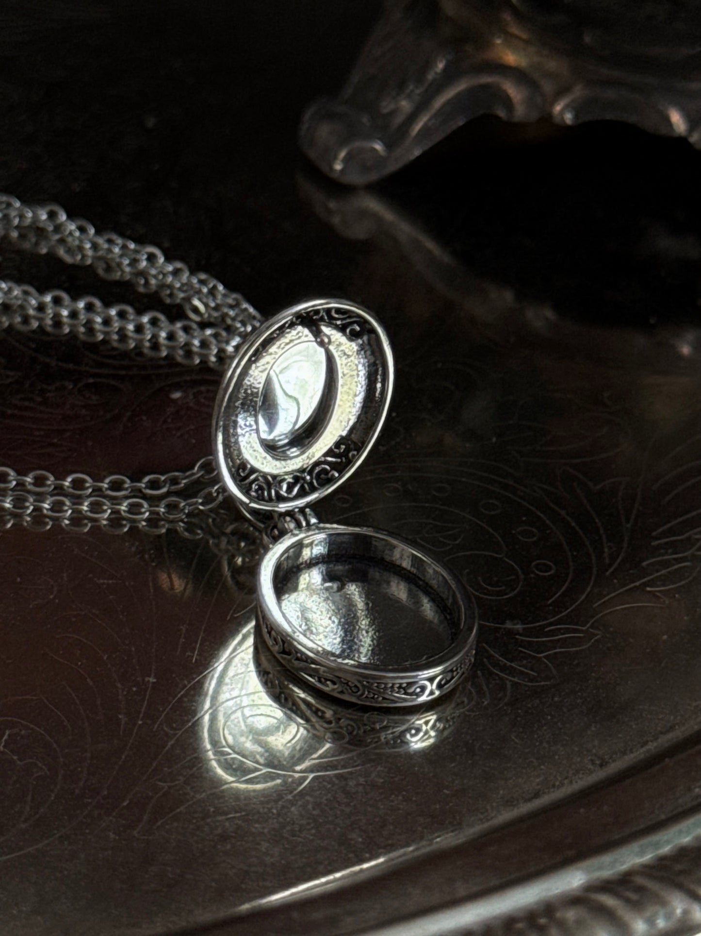 A victorian inspired silverwork locket necklace with filigree designs and a faux moonstone cabochon, pictured open on a vintage silver tray