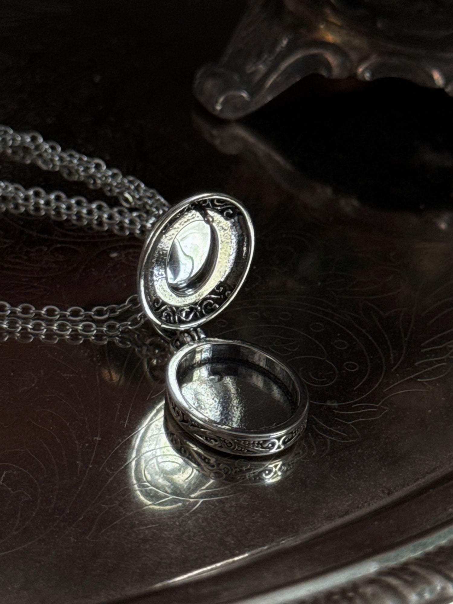 A victorian inspired silverwork locket necklace with filigree designs and a faux moonstone cabochon, pictured open on a vintage silver tray