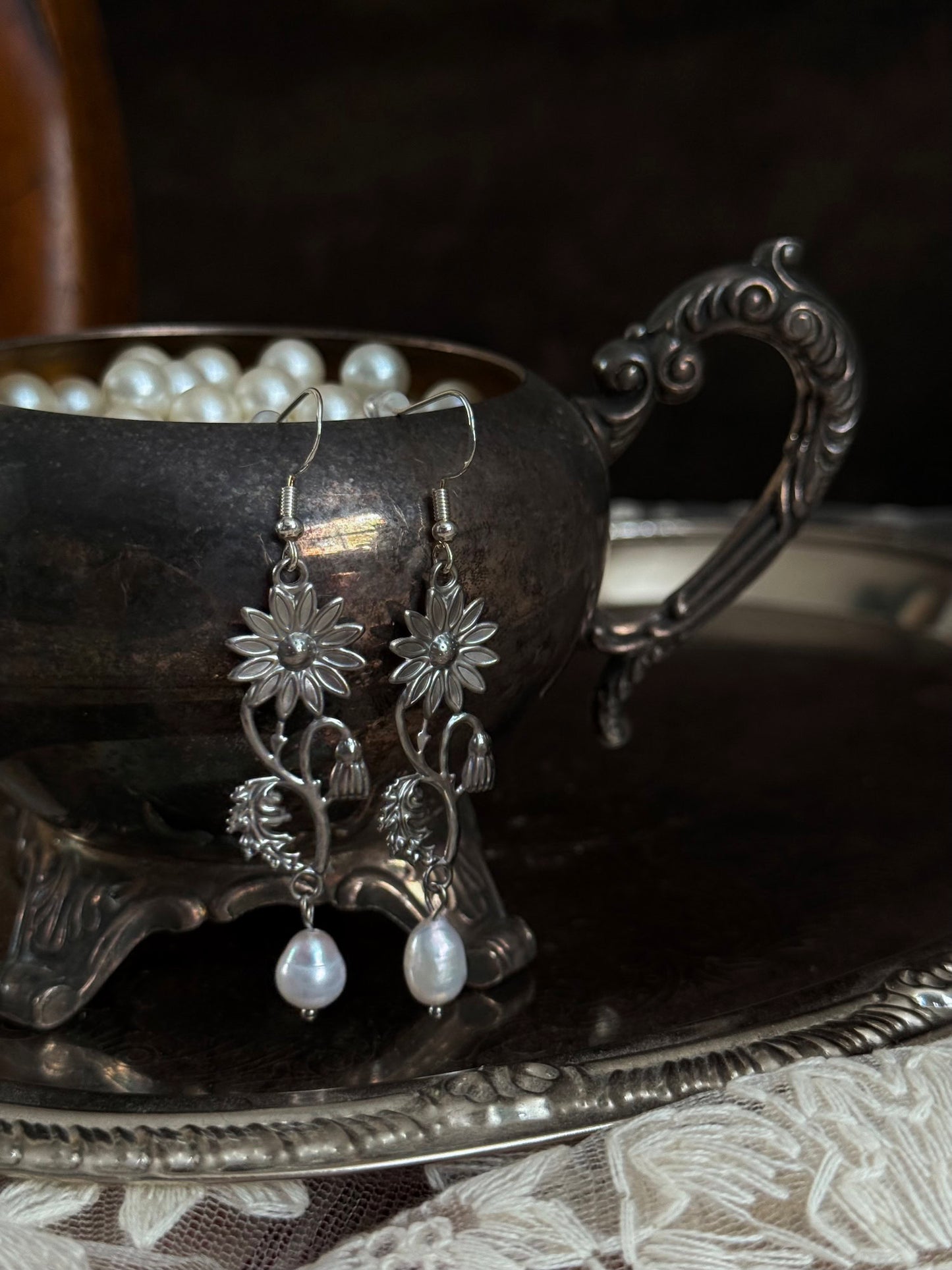 A pair of silver metal medieval wildflower daisy earrings with pearl drop dangles, hanging displayed on an antique silver dish.
