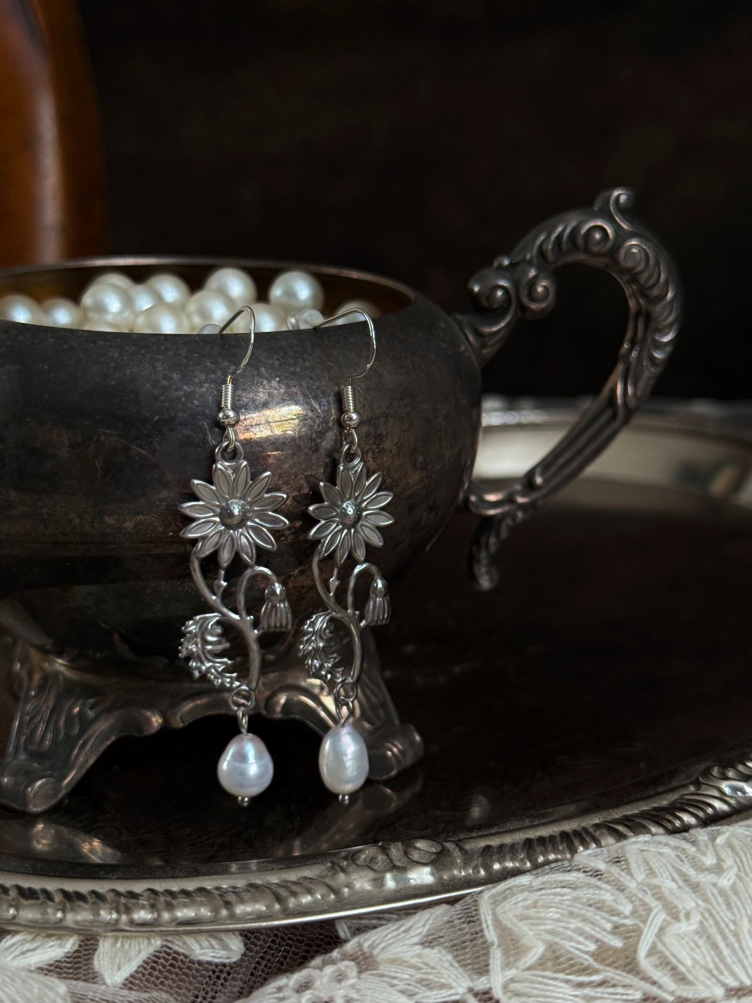 A pair of silver metal medieval wildflower daisy earrings with pearl drop dangles, hanging displayed on an antique silver dish.