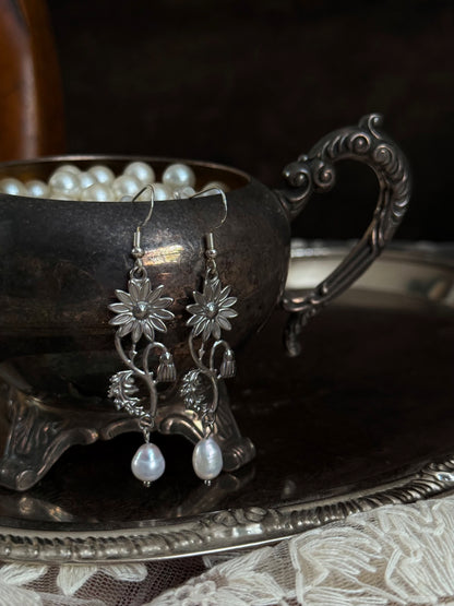 A pair of silver metal medieval wildflower daisy earrings with pearl drop dangles, hanging displayed on an antique silver dish.