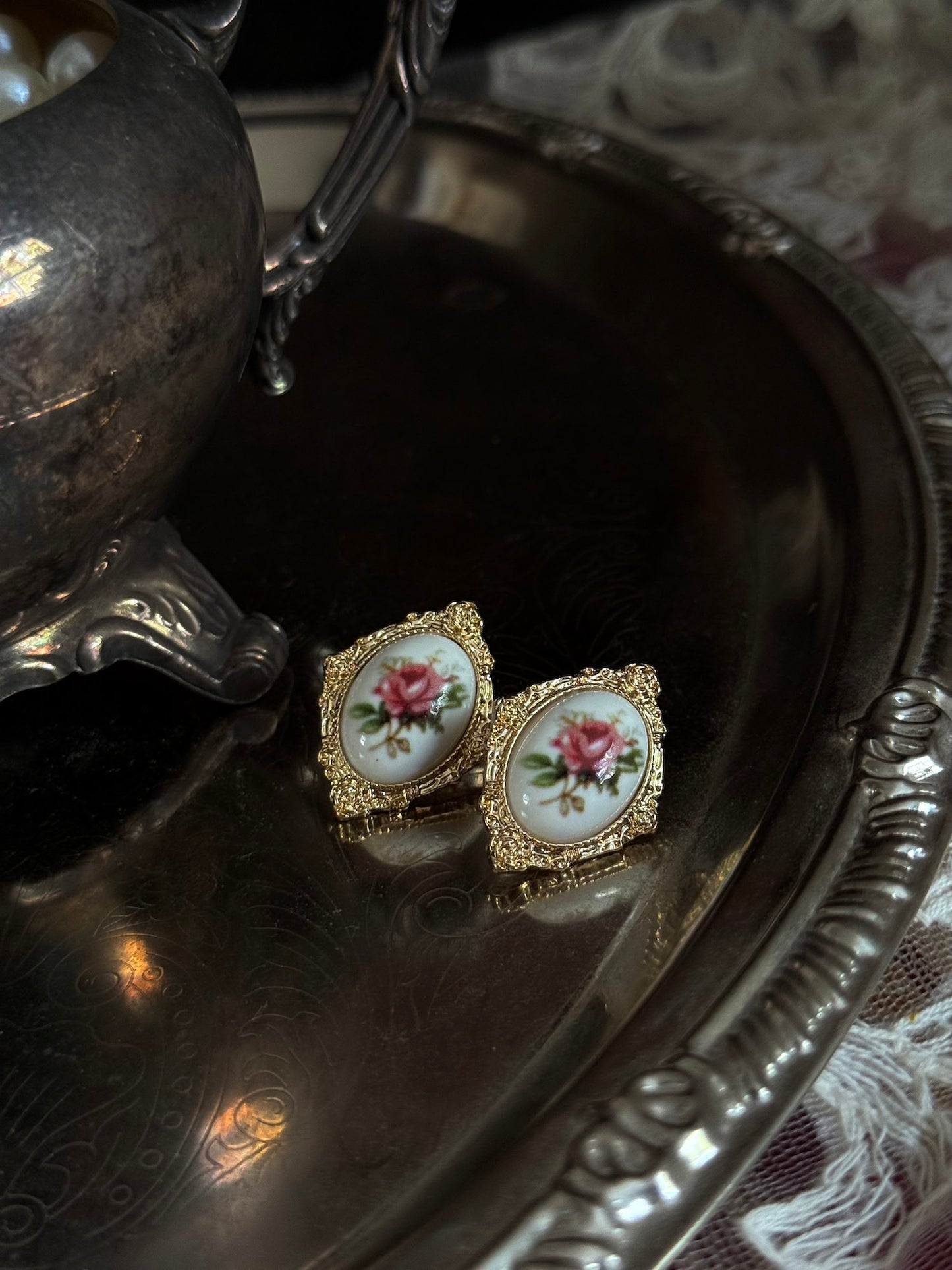 Pair of Historically-Inspired Painted Rose Ceramic gold-framed earrings displayed on an antique silver dish