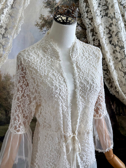 A medieval Fantasy Inspired Ivory Lace Bell Sleeve Tunic Duster with mesh accents is pictured on a mannequin in front of a painted woodland backdrop.