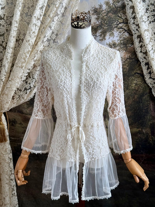 A medieval Fantasy Inspired Ivory Lace Bell Sleeve Tunic Duster with mesh accents is pictured on a mannequin in front of a painted woodland backdrop.