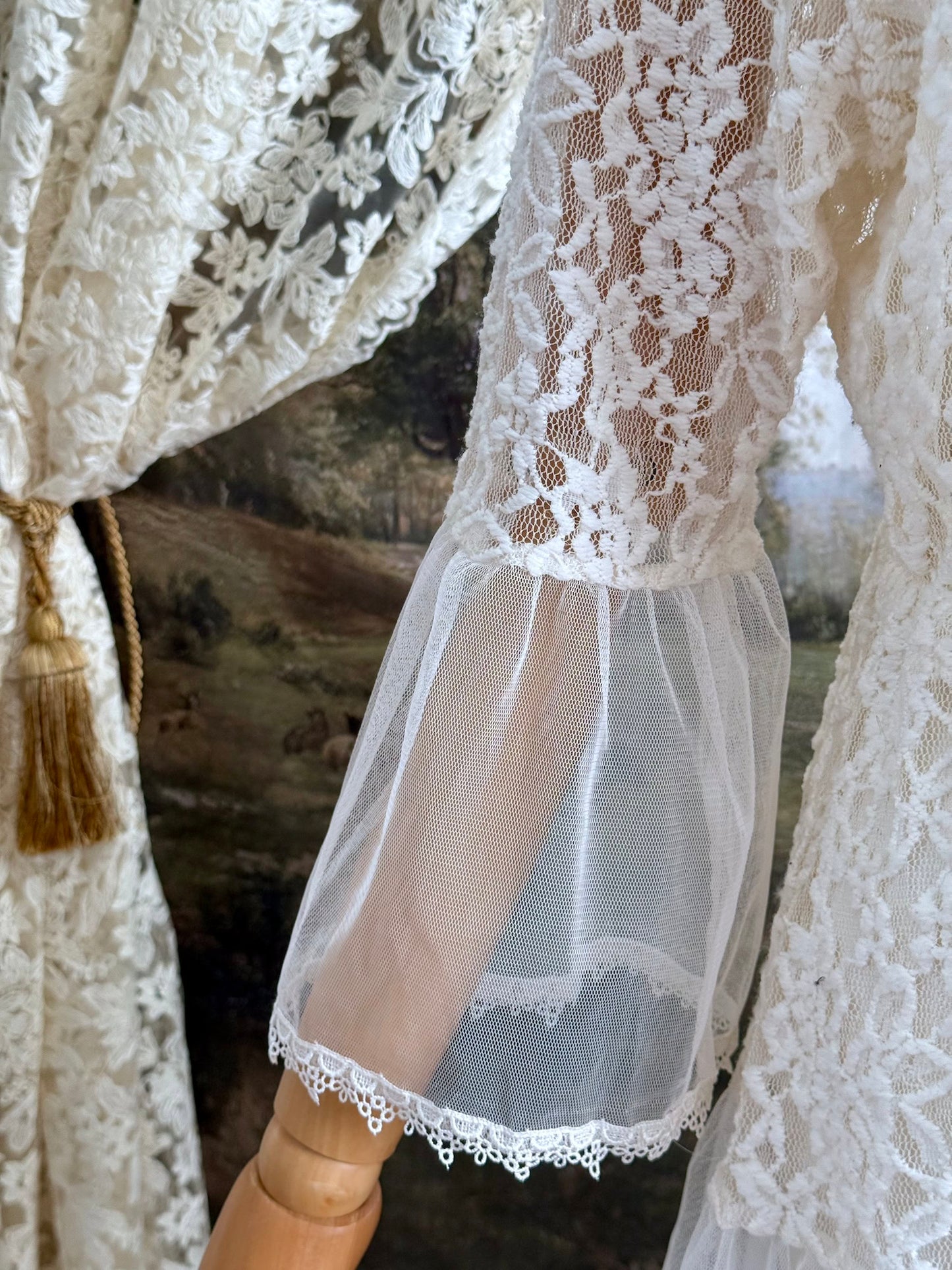 A medieval Fantasy Inspired Ivory Lace Bell Sleeve Tunic Duster with mesh accents is pictured on a mannequin in front of a painted woodland backdrop. Sleeve detail.