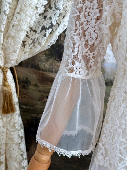 A medieval Fantasy Inspired Ivory Lace Bell Sleeve Tunic Duster with mesh accents is pictured on a mannequin in front of a painted woodland backdrop. Sleeve detail.