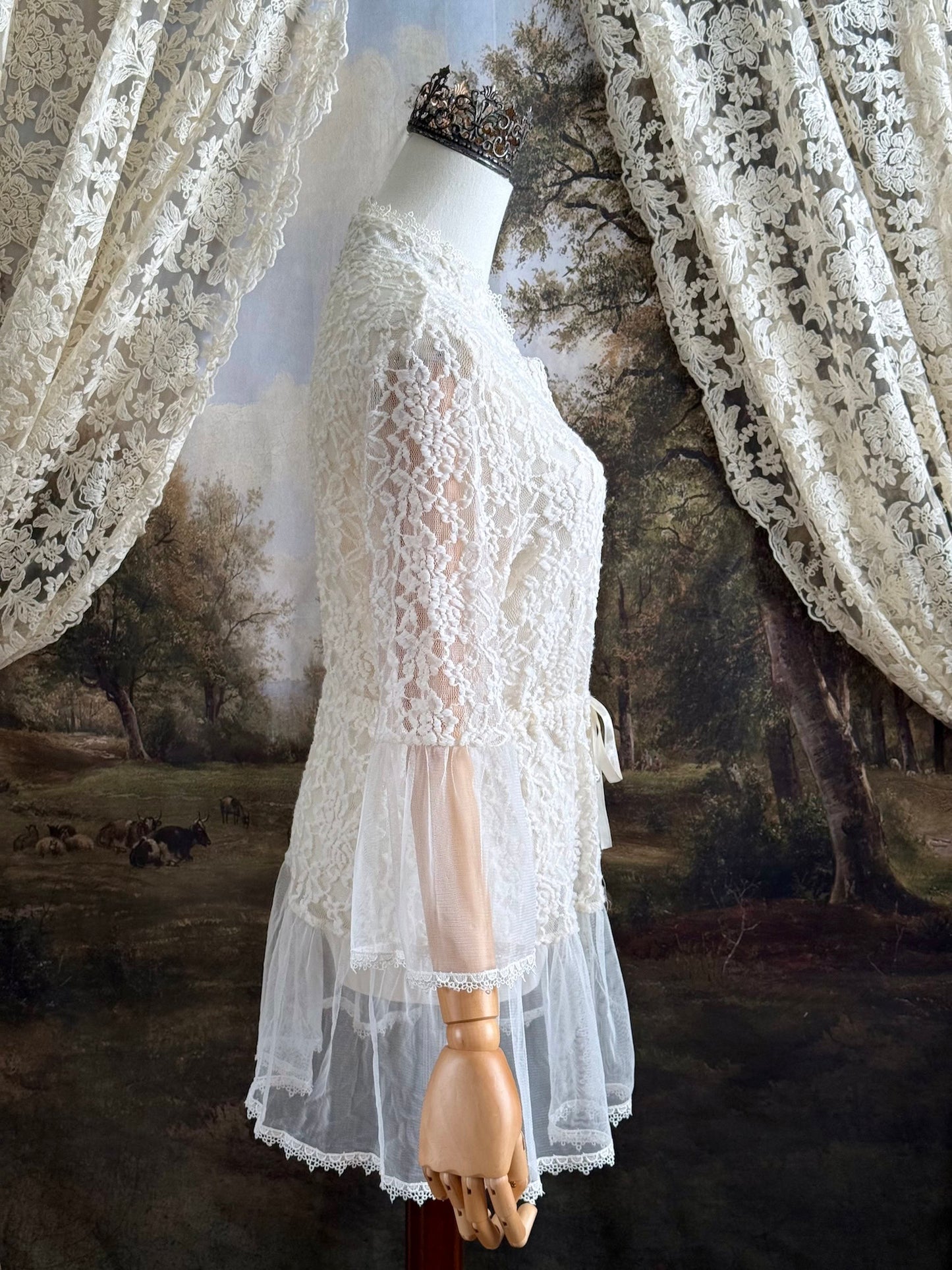 A medieval Fantasy Inspired Ivory Lace Bell Sleeve Tunic Duster with mesh accents is pictured on a mannequin in front of a painted woodland backdrop.