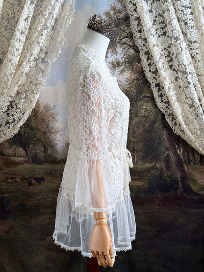 A medieval Fantasy Inspired Ivory Lace Bell Sleeve Tunic Duster with mesh accents is pictured on a mannequin in front of a painted woodland backdrop.