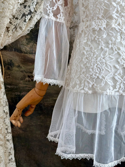 A medieval Fantasy Inspired Ivory Lace Bell Sleeve Tunic Duster with mesh accents is pictured on a mannequin in front of a painted woodland backdrop.