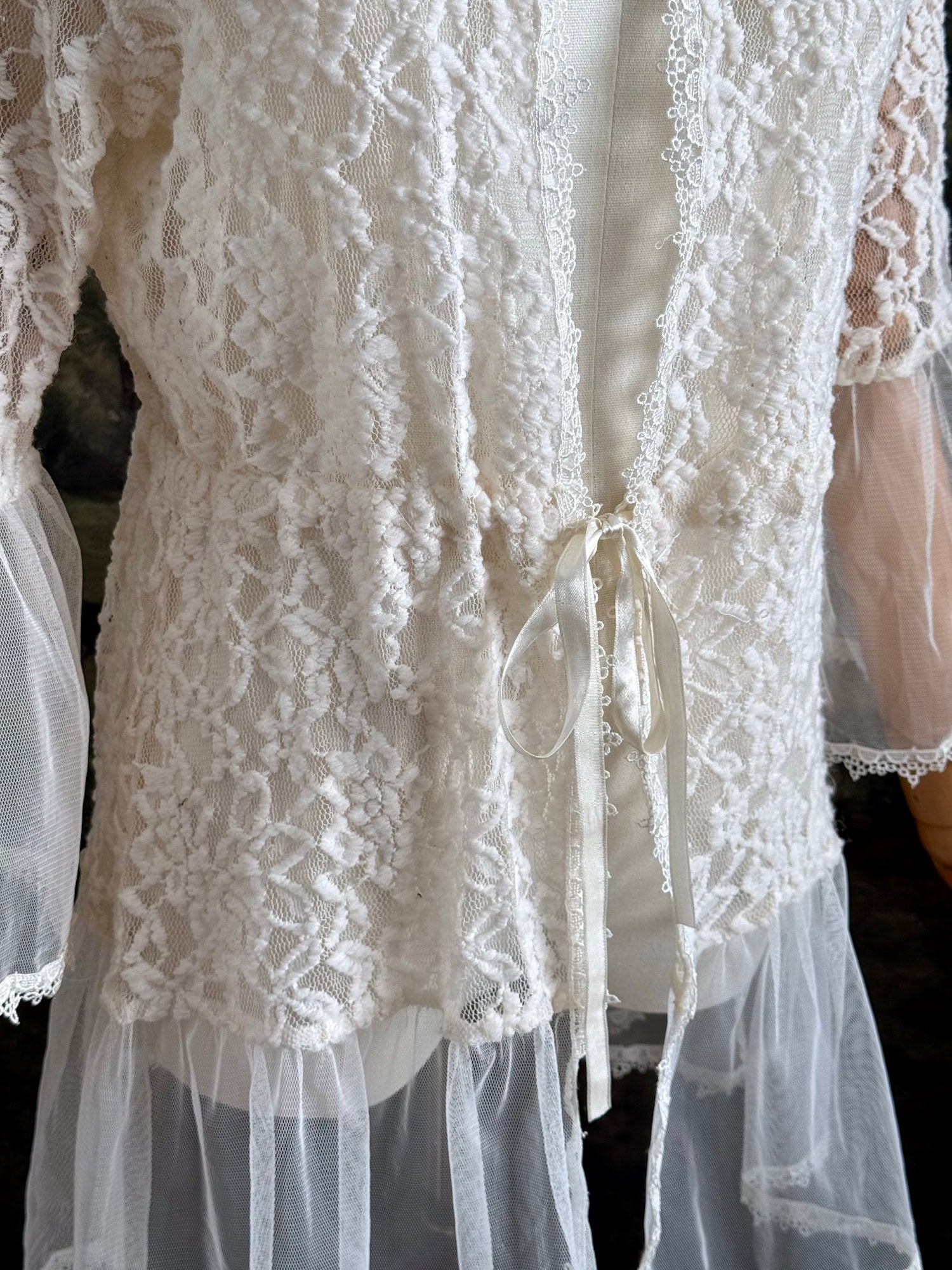 A medieval Fantasy Inspired Ivory Lace Bell Sleeve Tunic Duster with mesh accents is pictured on a mannequin in front of a painted woodland backdrop. Detail of ribbon.