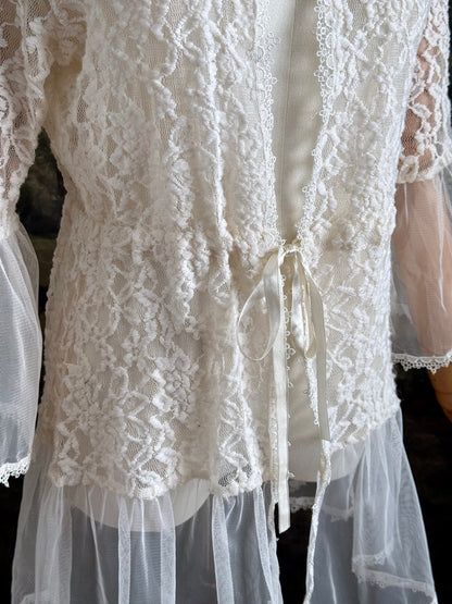 A medieval Fantasy Inspired Ivory Lace Bell Sleeve Tunic Duster with mesh accents is pictured on a mannequin in front of a painted woodland backdrop. Detail of ribbon.