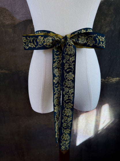A medieval and renaissance inspired dark teal green silk ribbon belt with an ornate gold damask pattern and gold engraved belt tips, tied in a bow around a mannequin in front of a historical painting backdrop.