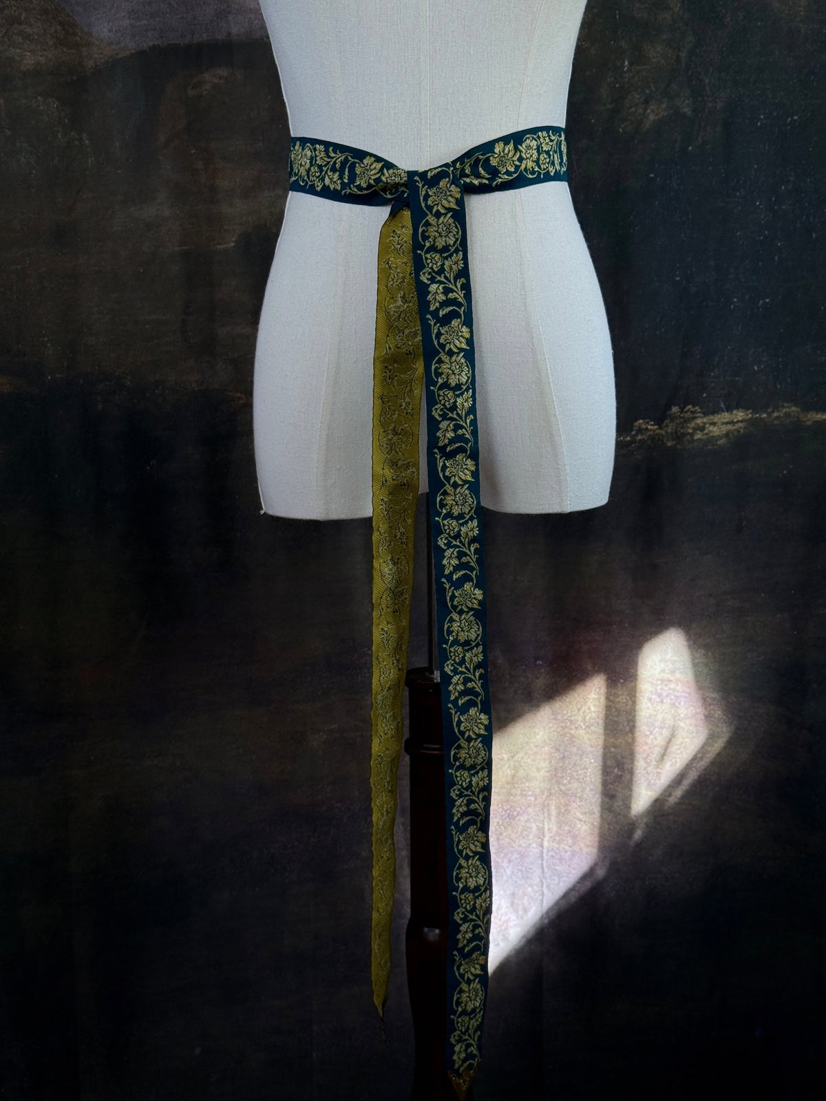A medieval and renaissance inspired dark teal green silk ribbon belt with an ornate gold damask pattern and gold engraved belt tips, tied in a bow around a mannequin in front of a historical painting backdrop.