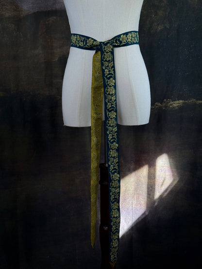 A medieval and renaissance inspired dark teal green silk ribbon belt with an ornate gold damask pattern and gold engraved belt tips, tied in a bow around a mannequin in front of a historical painting backdrop.