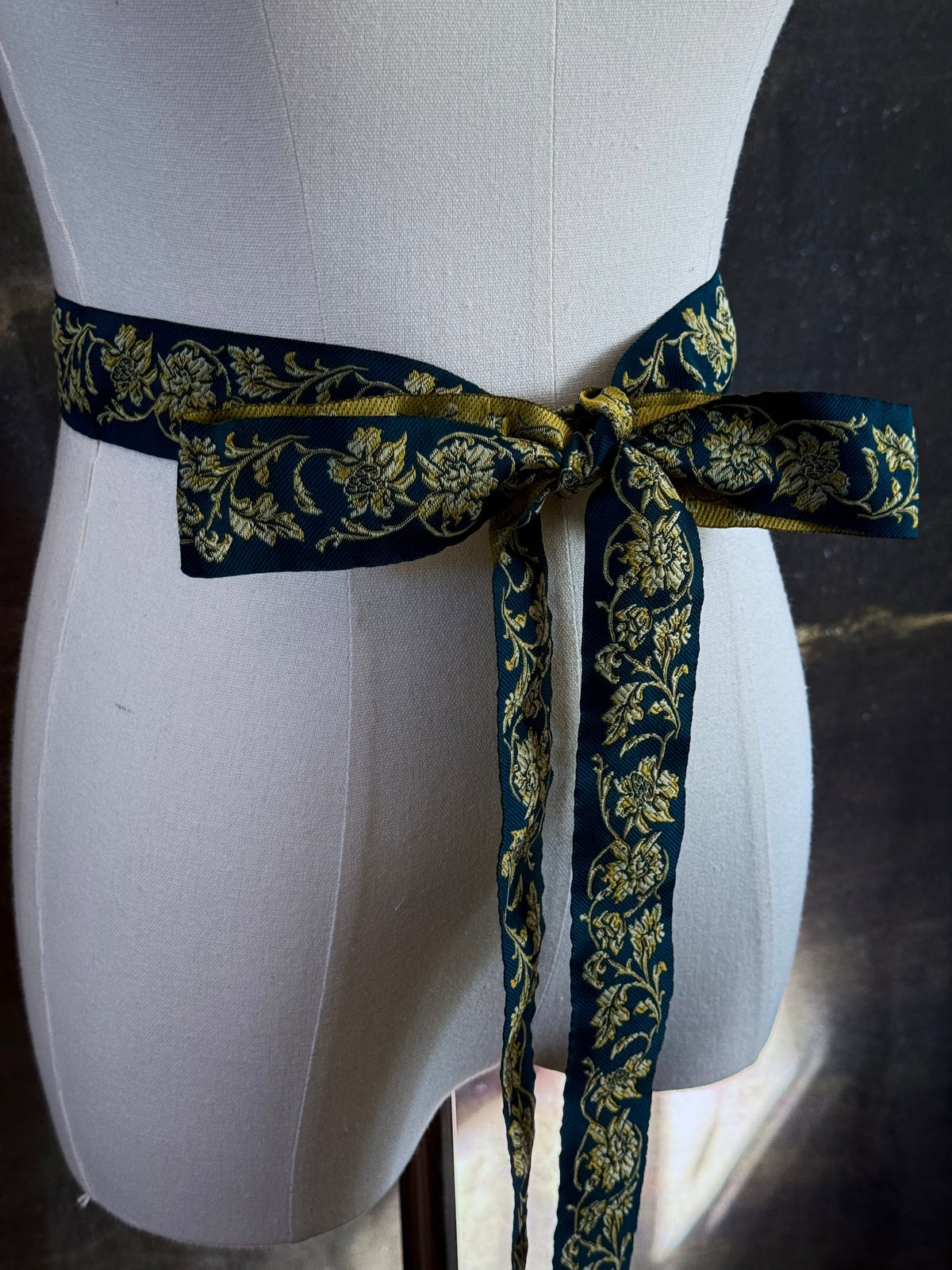 A medieval and renaissance inspired dark teal green silk ribbon belt with an ornate gold damask pattern and gold engraved belt tips, tied in a bow around a mannequin in front of a historical painting backdrop.