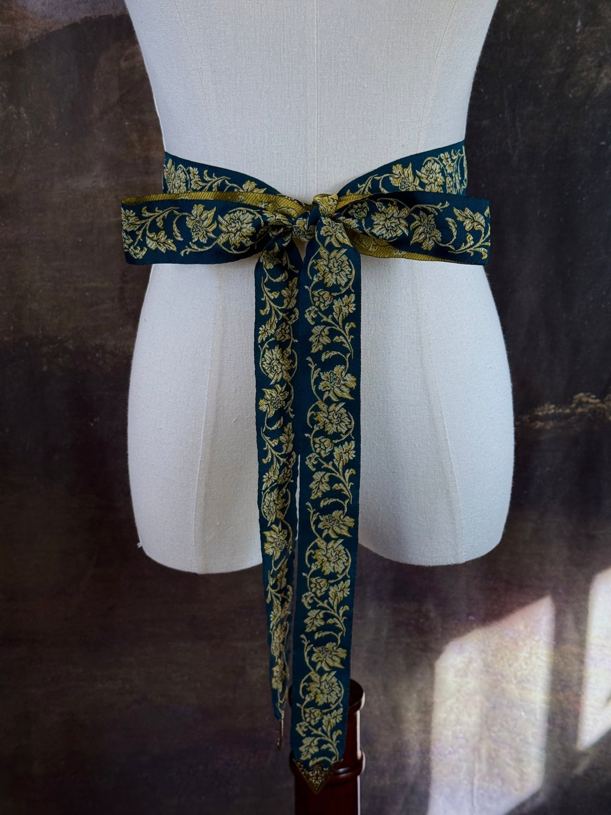A medieval and renaissance inspired dark teal green silk ribbon belt with an ornate gold damask pattern and gold engraved belt tips, tied in a bow around a mannequin in front of a historical painting backdrop.