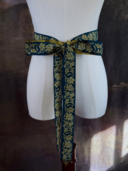 A medieval and renaissance inspired dark teal green silk ribbon belt with an ornate gold damask pattern and gold engraved belt tips, tied in a bow around a mannequin in front of a historical painting backdrop.