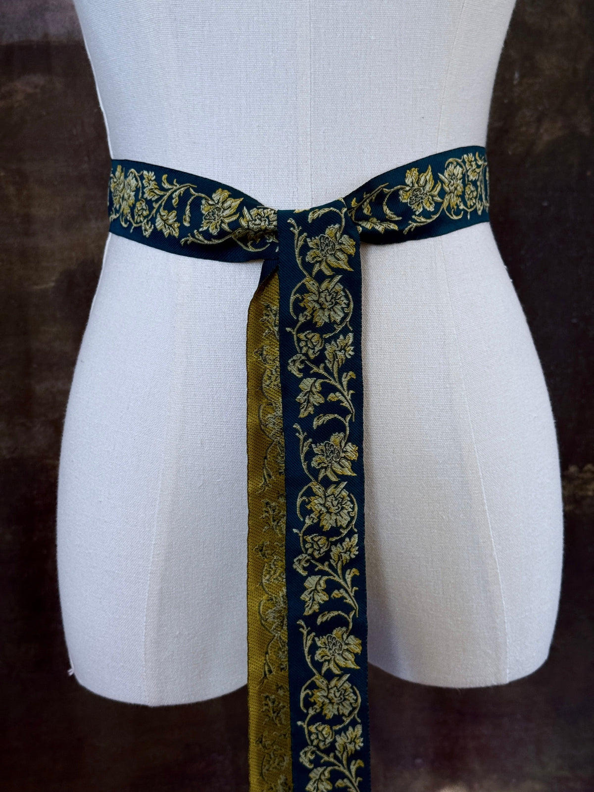 A medieval and renaissance inspired dark teal green silk ribbon belt with an ornate gold damask pattern and gold engraved belt tips, tied in a bow around a mannequin in front of a historical painting backdrop.