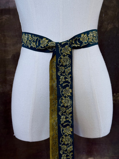 A medieval and renaissance inspired dark teal green silk ribbon belt with an ornate gold damask pattern and gold engraved belt tips, tied in a bow around a mannequin in front of a historical painting backdrop.