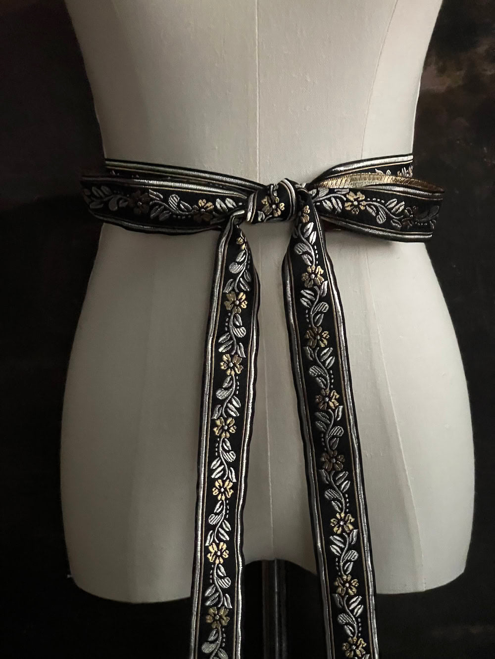 An ornate black medieval ribbon belt with silver and gold metallic floral embroidery woven into it, tied into a bow on a mannequin in front of an art history backdrop.