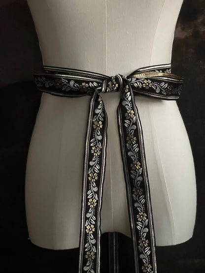 An ornate black medieval ribbon belt with silver and gold metallic floral embroidery woven into it, tied into a bow on a mannequin in front of an art history backdrop.