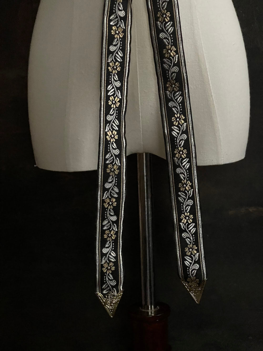 An ornate black medieval ribbon belt with silver and gold metallic floral embroidery woven into it, tied into a bow on a mannequin in front of an art history backdrop.