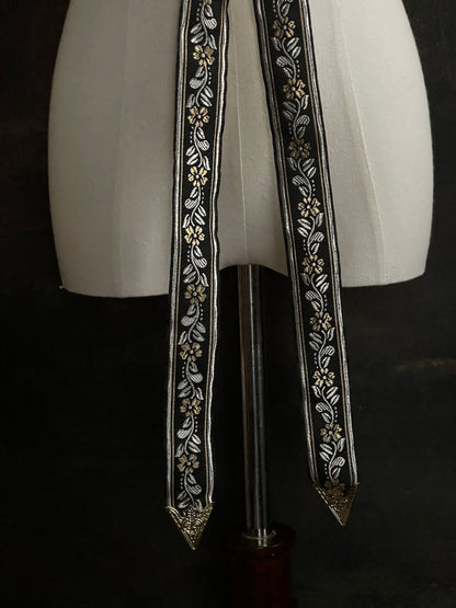 An ornate black medieval ribbon belt with silver and gold metallic floral embroidery woven into it, tied into a bow on a mannequin in front of an art history backdrop.