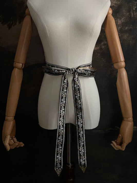 An ornate black medieval ribbon belt with silver and gold metallic floral embroidery woven into it, tied into a bow on a mannequin in front of an art history backdrop.