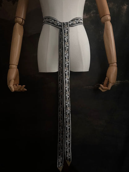 An ornate black medieval ribbon belt with silver and gold metallic floral embroidery woven into it, on a mannequin in front of an art history backdrop.