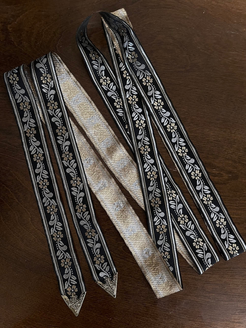 An ornate black medieval ribbon belt with silver and gold metallic floral embroidery woven into it, on a wooden table
