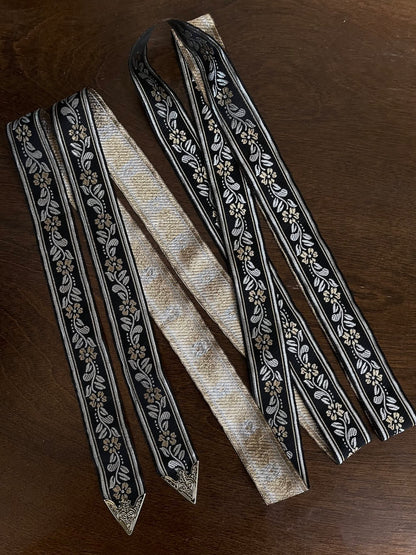 An ornate black medieval ribbon belt with silver and gold metallic floral embroidery woven into it, on a wooden table