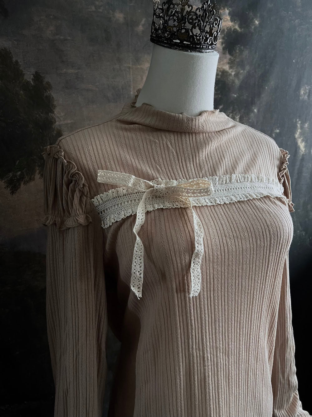 A Beige / light brown Victorian Inspired Ribbed Knit Long-Sleeve Top with ribbon Bow Applique and ruffle trim in front of an art history painting backdrop
