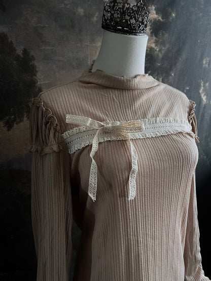 A Beige / light brown Victorian Inspired Ribbed Knit Long-Sleeve Top with ribbon Bow Applique and ruffle trim in front of an art history painting backdrop