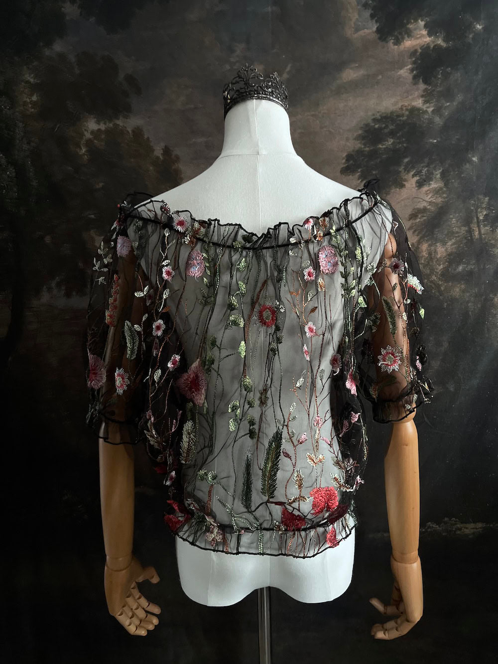 Mannequin wearing a gothic black sheer mesh puff sleeve blouse with embroidered botanicals and a ruffle hem, on a forest-themed art history background
