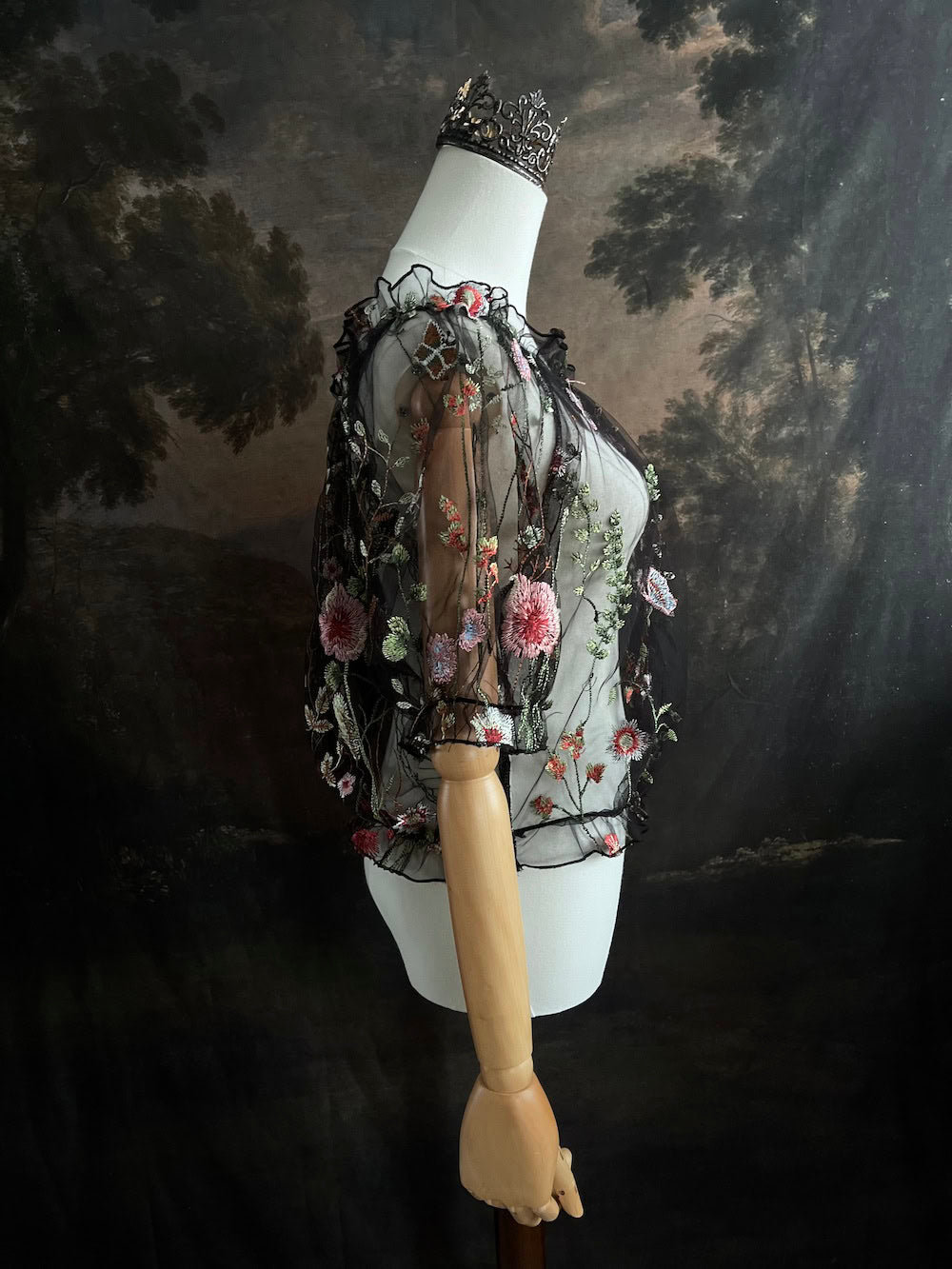 Mannequin wearing a gothic black sheer mesh puff sleeve blouse with embroidered botanicals and a ruffle hem, on a forest-themed art history background