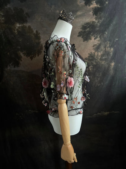 Mannequin wearing a gothic black sheer mesh puff sleeve blouse with embroidered botanicals and a ruffle hem, on a forest-themed art history background
